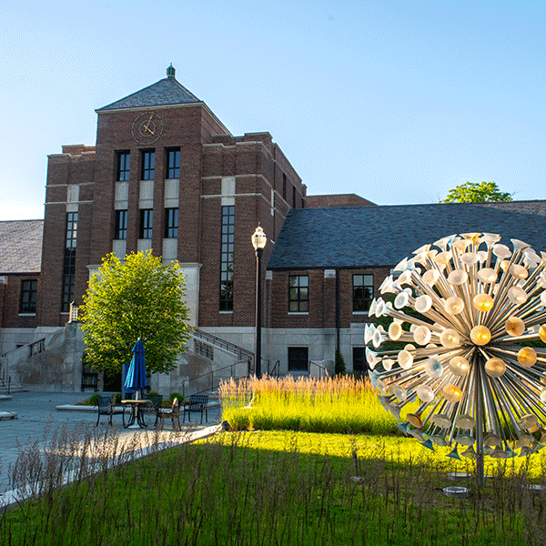 Outdoor scene featuring a modern metal sculpture resembling a dandelion, with a brick building in the background adorned with a clock tower. The setting includes landscaping with green grass, shrubs, and a tree, as well as outdoor seating shaded by blue umbrellas under a clear blue sky